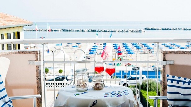A balcony with a table set for two, overlooking a beach with umbrellas and flags. - Hotel Alsen (Rimini)