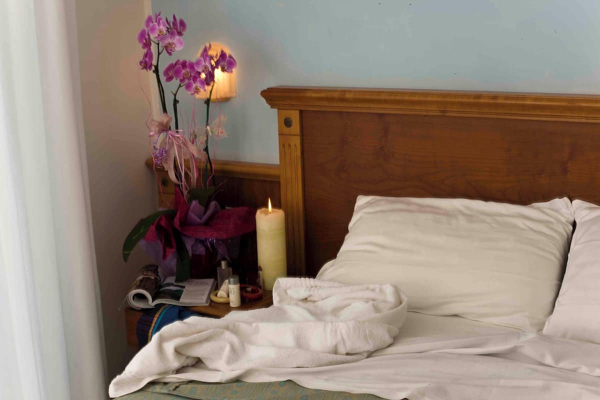 A neatly made bed with a white blanket, a wooden headboard, and a bedside table with a lit candle, a vase of flowers, and some personal items.