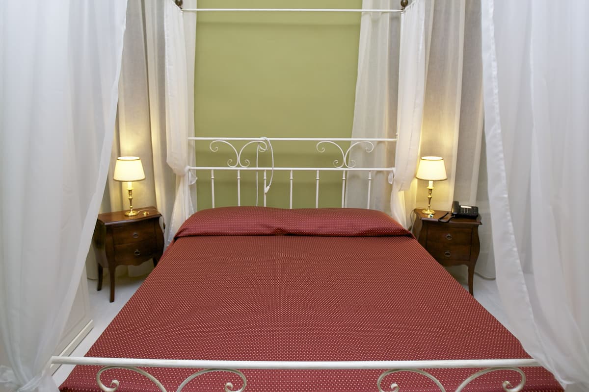 Una cama individual con estructura metálica, cortinas blancas y dos mesitas de noche con lámparas.