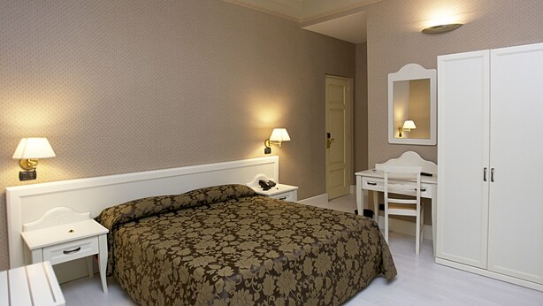 A hotel room with a large bed, a desk, a chair, a wardrobe, and two bedside lamps. - Palazzo Riario (Viterbo)