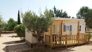 Una pequeña casa móvil de un piso con una terraza de madera y un árbol enfrente.