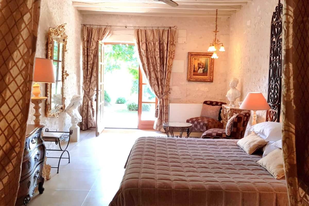 Een slaapkamer met een groot bed, een kaptafel, een stoel en een tafel. Er is een raam met gordijnen en uitzicht op groen.