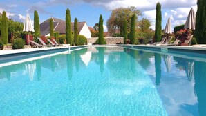 Piscina azul cristalina rodeada por espreguiçadeiras e guarda-sóis, com uma parede de pedra e árvores ao fundo.