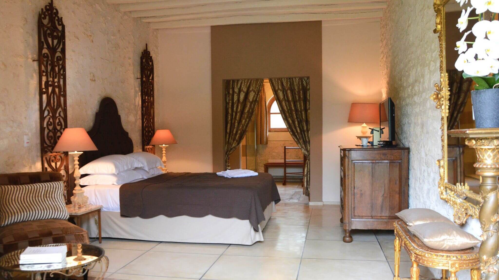 A room with a bed, two bedside tables with lamps, a chair, a small table, and a mirror with flowers.