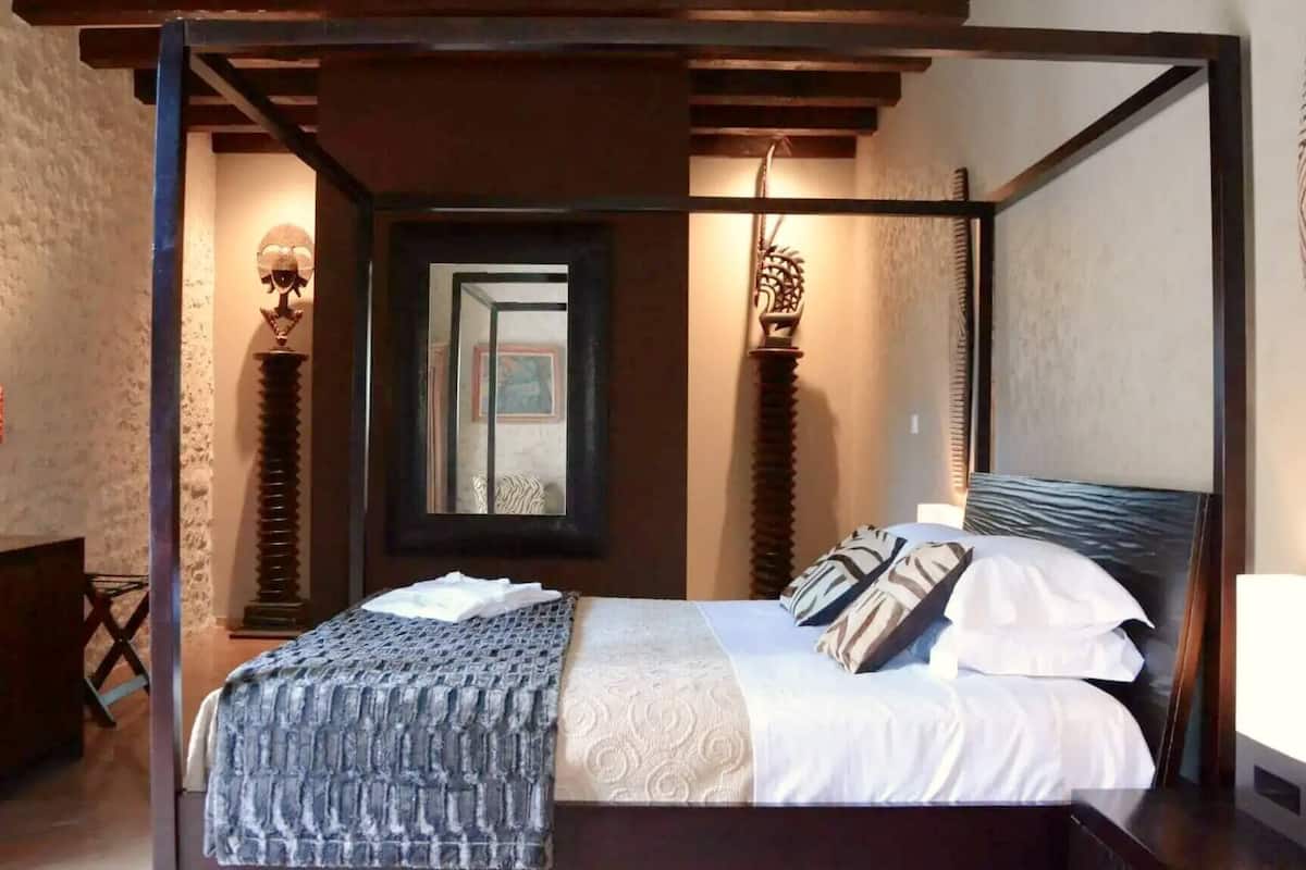 Een slaapkamer met een hemelbed, een houten hoofdbord, een nachtkastje, een lamp en een spiegel.