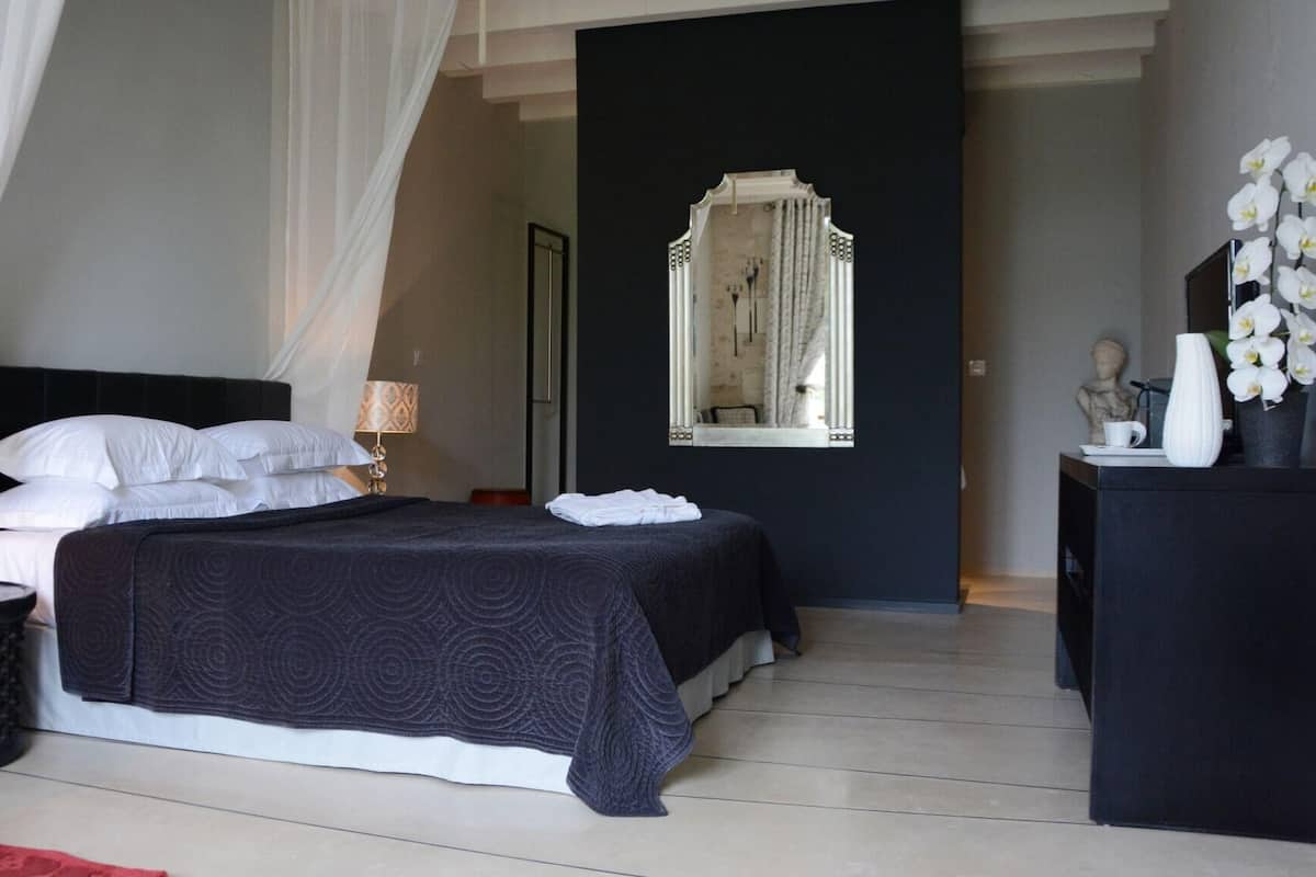 Een slaapkamer met een bed, nachtkastje, lamp, spiegel en een dressoir met decoratieve objecten.