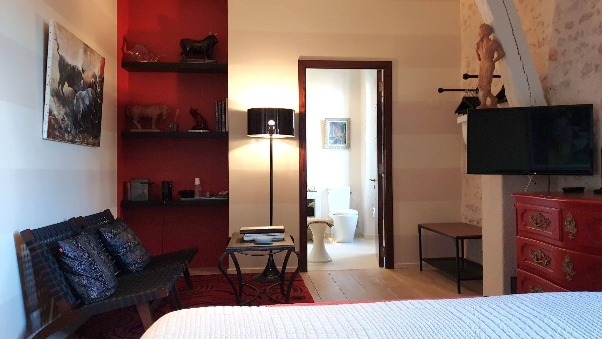 A hotel room with a bed, a desk, a chair, a TV, and a red wall with shelves.