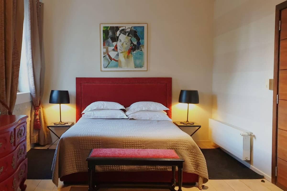 Een hotelkamer met een rode hoofdbord, een bed met witte lakens, twee nachtkastjes met lampen, een rode bank, een dressoir en een schilderij aan de muur.