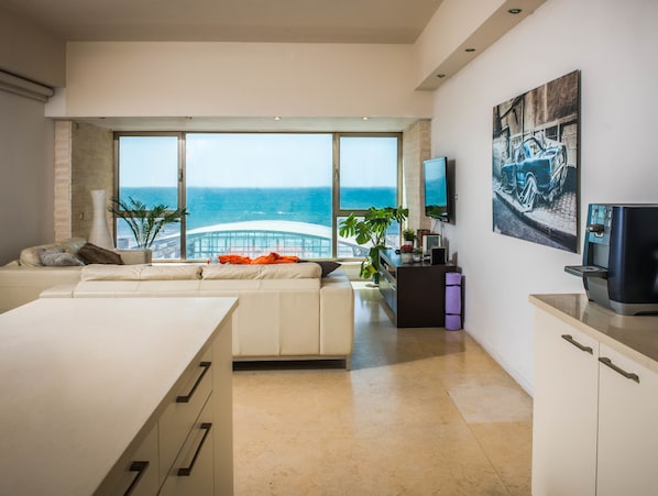 A modern living room with a large window overlooking the ocean, a sofa, a coffee machine, and a flat-screen TV on the wall.