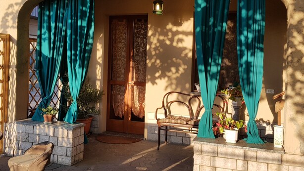 Una entrada exterior con puerta de madera, cortinas color turquesa, plantas en macetas y una silla de mimbre.