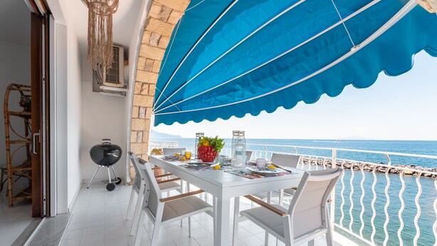 A balcony with a white table set for dining, a grill, and a view of the sea. - Villa del Sole (Massa Lubrense)