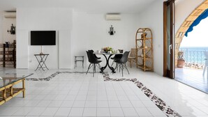A modern living room with a dining area, a TV mounted on the wall, and a view of the sea through an open door. - Villa del Sole (Massa Lubrense)
