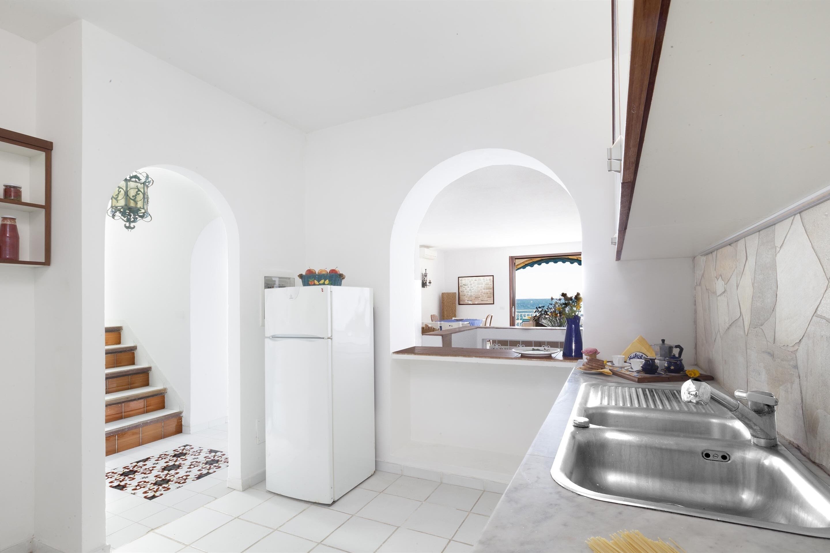 A modern kitchen with white walls, a stainless steel sink, and a refrigerator. There is a wooden staircase leading to another room.