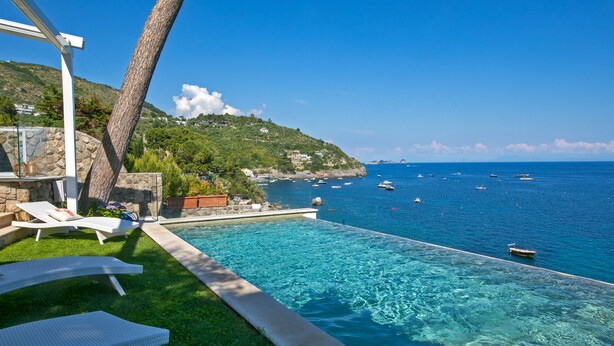 Zona junto a la piscina con sillones de descanso, pared de piedra y vista al mar con barcos.