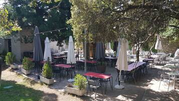 Un área de café al aire libre con mesas y sillas, algunas cubiertas por sombrillas, y plantas en macetas.
