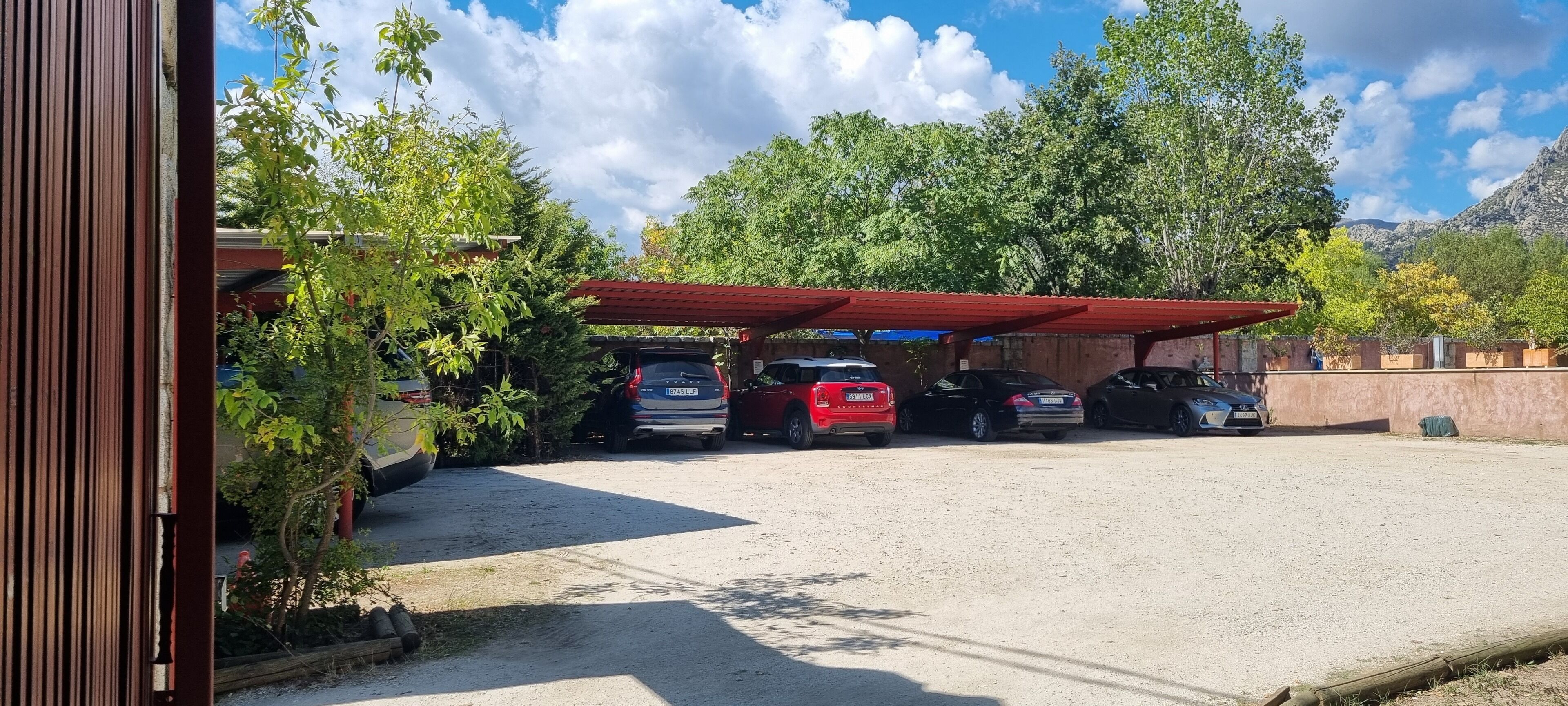 Un estacionamiento con varios coches bajo una cubierta roja, rodeado de vegetación y montañas al fondo.