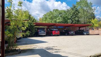 Un estacionamiento con varios coches bajo una cubierta roja, rodeado de vegetación y montañas al fondo.