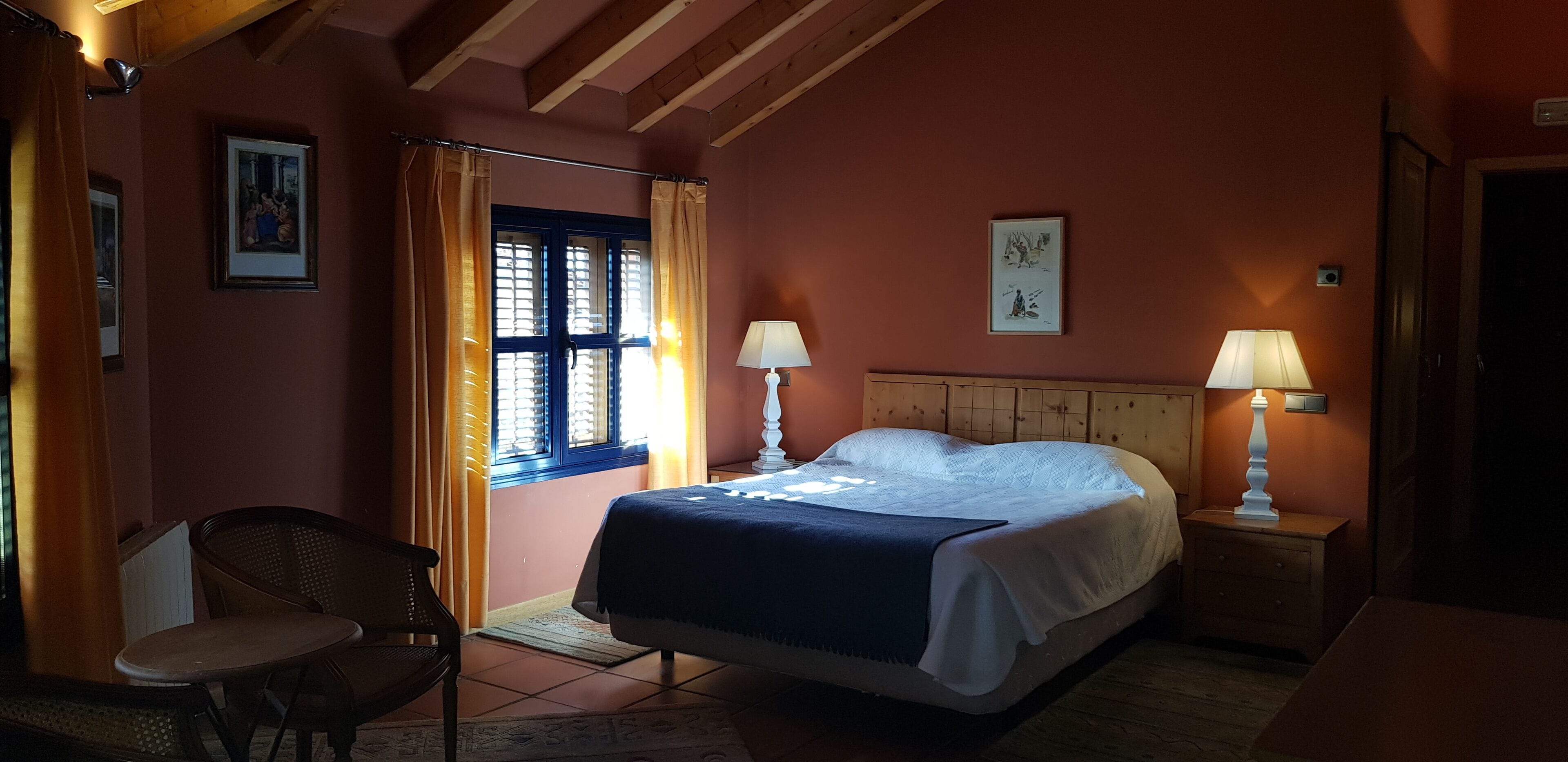 Ein Schlafzimmer mit einem Bett, zwei Nachttischlampen, einem Sessel, einem Fenster mit Vorhängen und einem gerahmten Bild an der Wand.