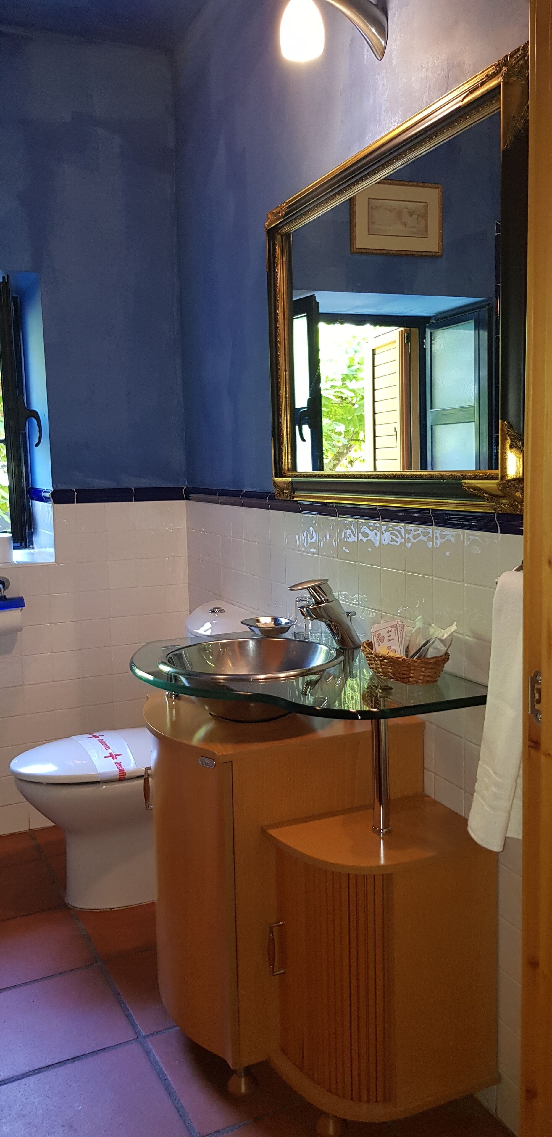 Un baño con lavamanos de vidrio, mueble de madera y espejo con marco dorado.