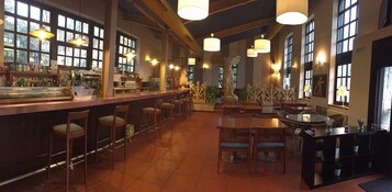 Zona de bar con mostrador, taburetes y mesas. Amplias ventanas y lámparas colgantes.
