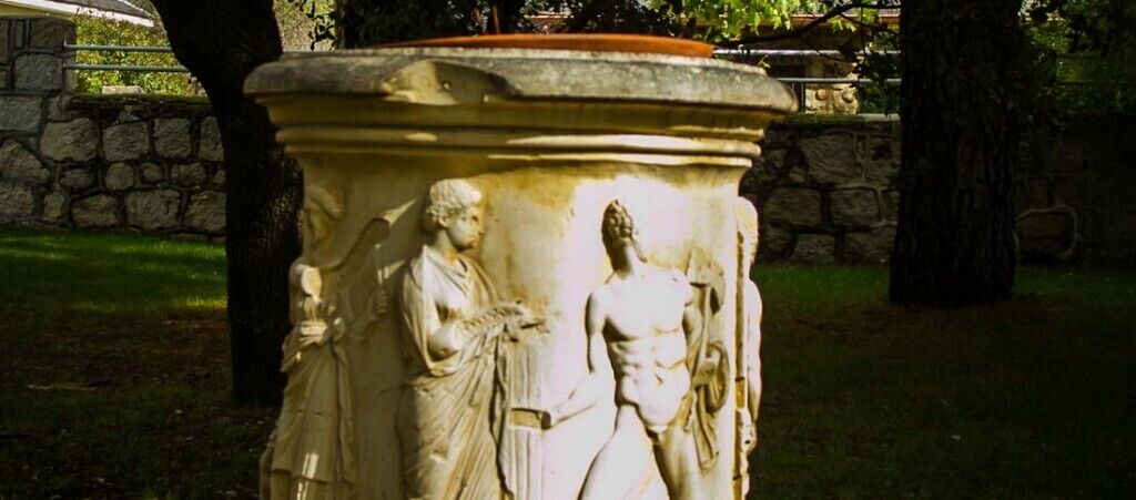 A stone pedestal with classical sculptures, surrounded by grass and trees.