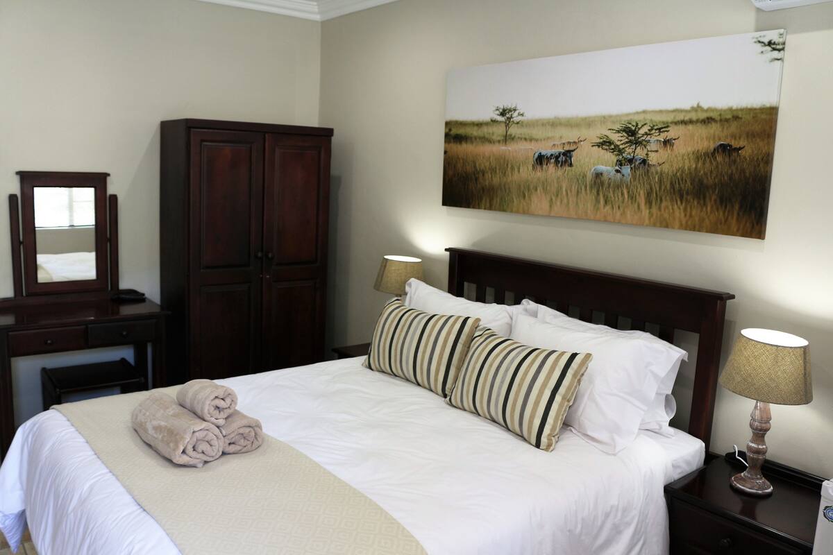 Ein ordentlich gemachtes Bett mit Kissen und Decke, ein Holzschrank, ein Schreibtisch mit Spiegel und ein an der Wand hängendes Gemälde einer Savannenlandschaft.