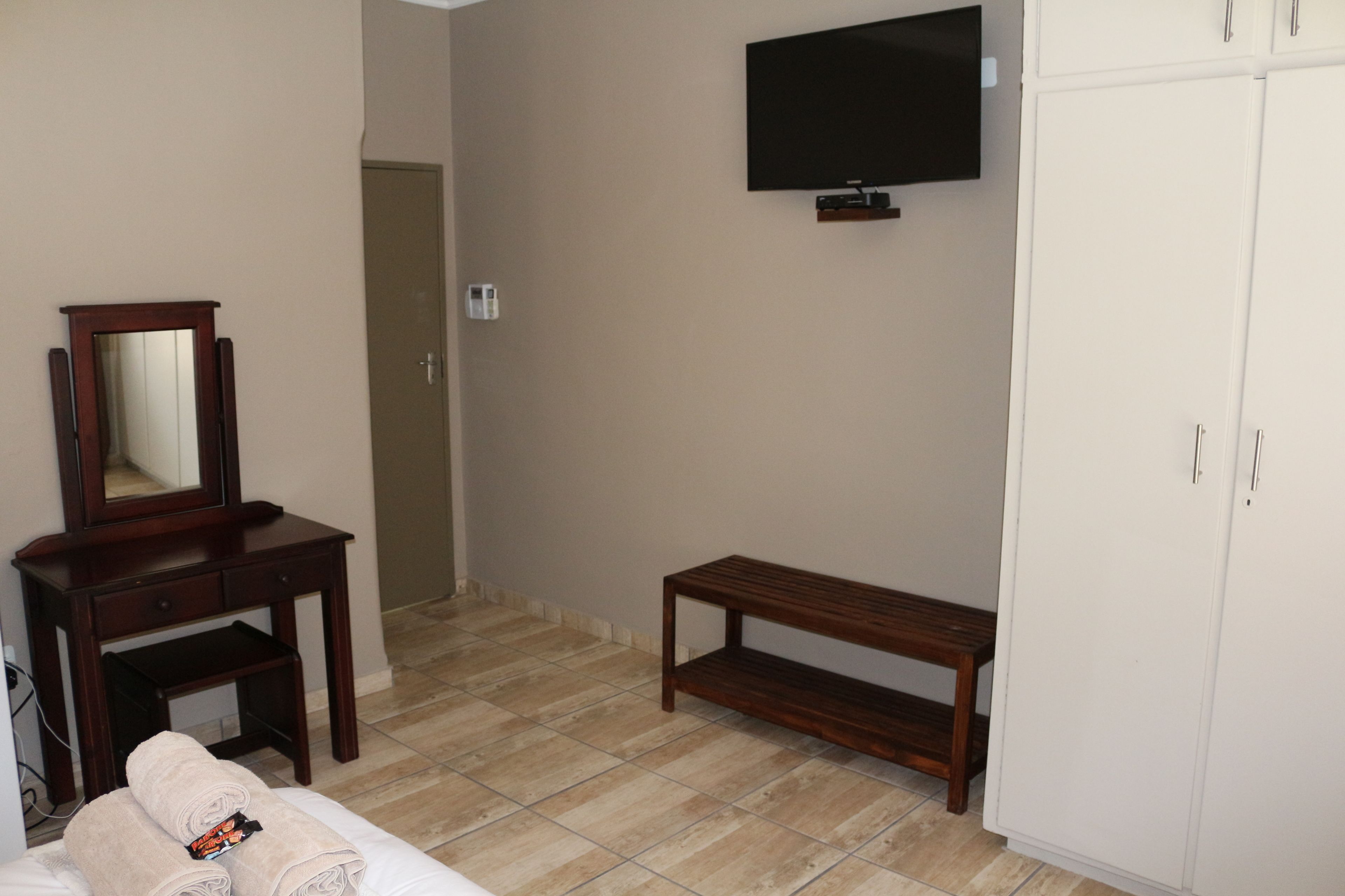 A hotel room with a bed, a wooden bench, a wardrobe, a mirror, and a television mounted on the wall.