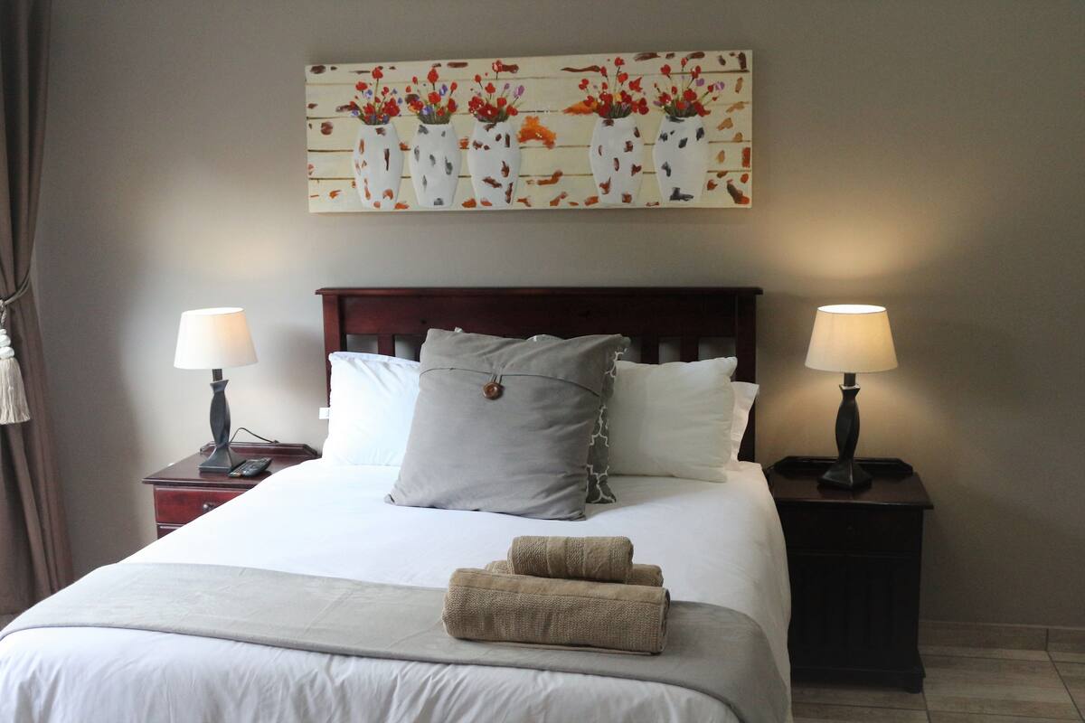 Ein ordentlich bezogenes Bett mit einem Kopfkissen, zwei Nachttischen mit Lampen und einem floralen Bild darüber.