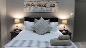 A neatly made bed with a patterned pillow, two bedside lamps, a decorative wall piece, and a folded towel on the bed. - Hummingbirds Nest Guesthouse (Mbombela)