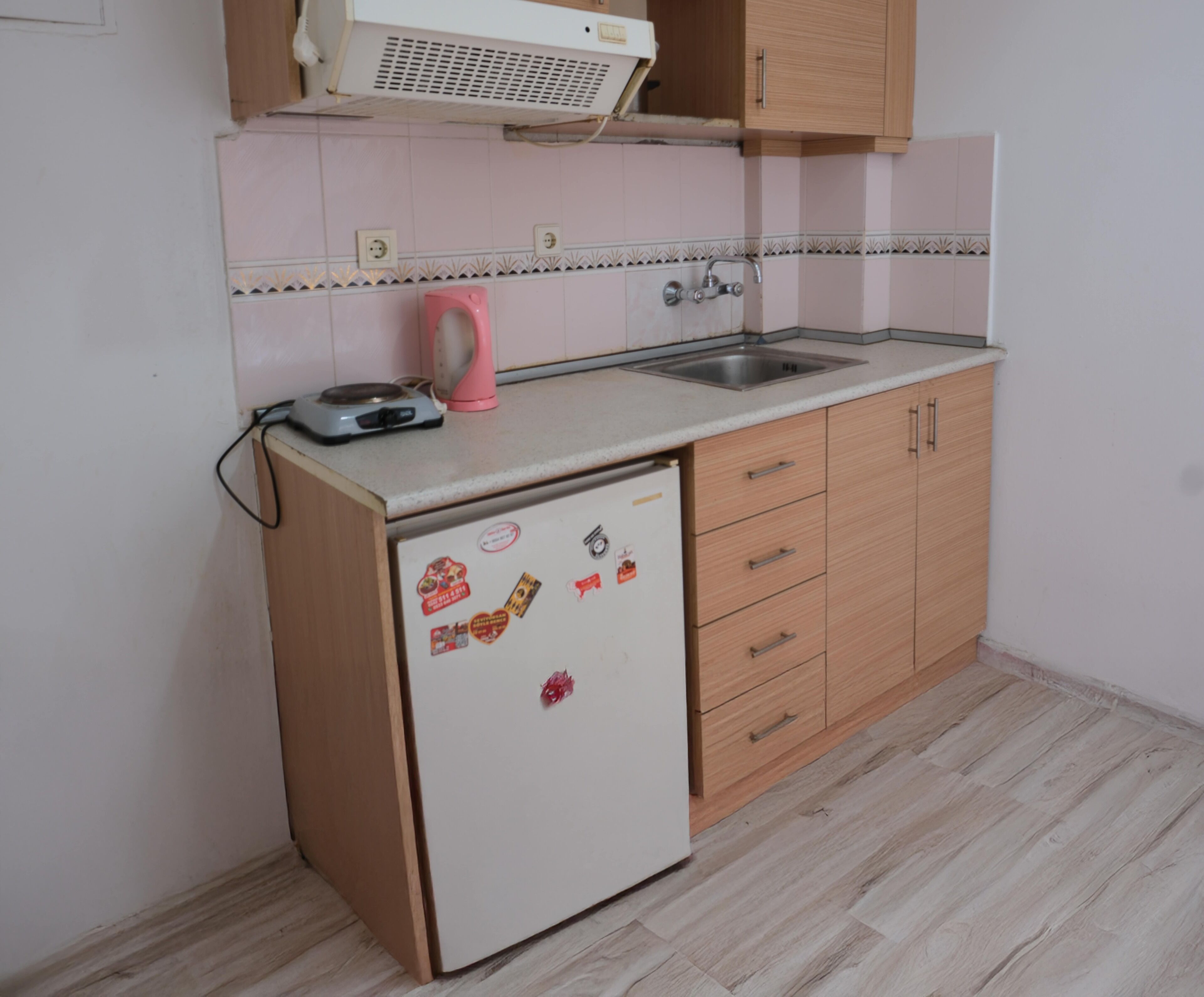 Een kleine keuken met een witte koelkast, houten kastjes, een spoelbak en een roze waterkoker.