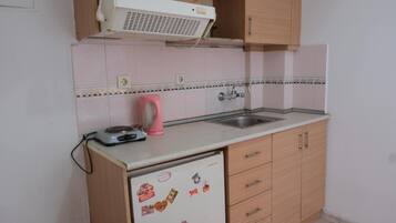 Een kleine keuken met een witte koelkast, houten kastjes, een spoelbak en een roze waterkoker.