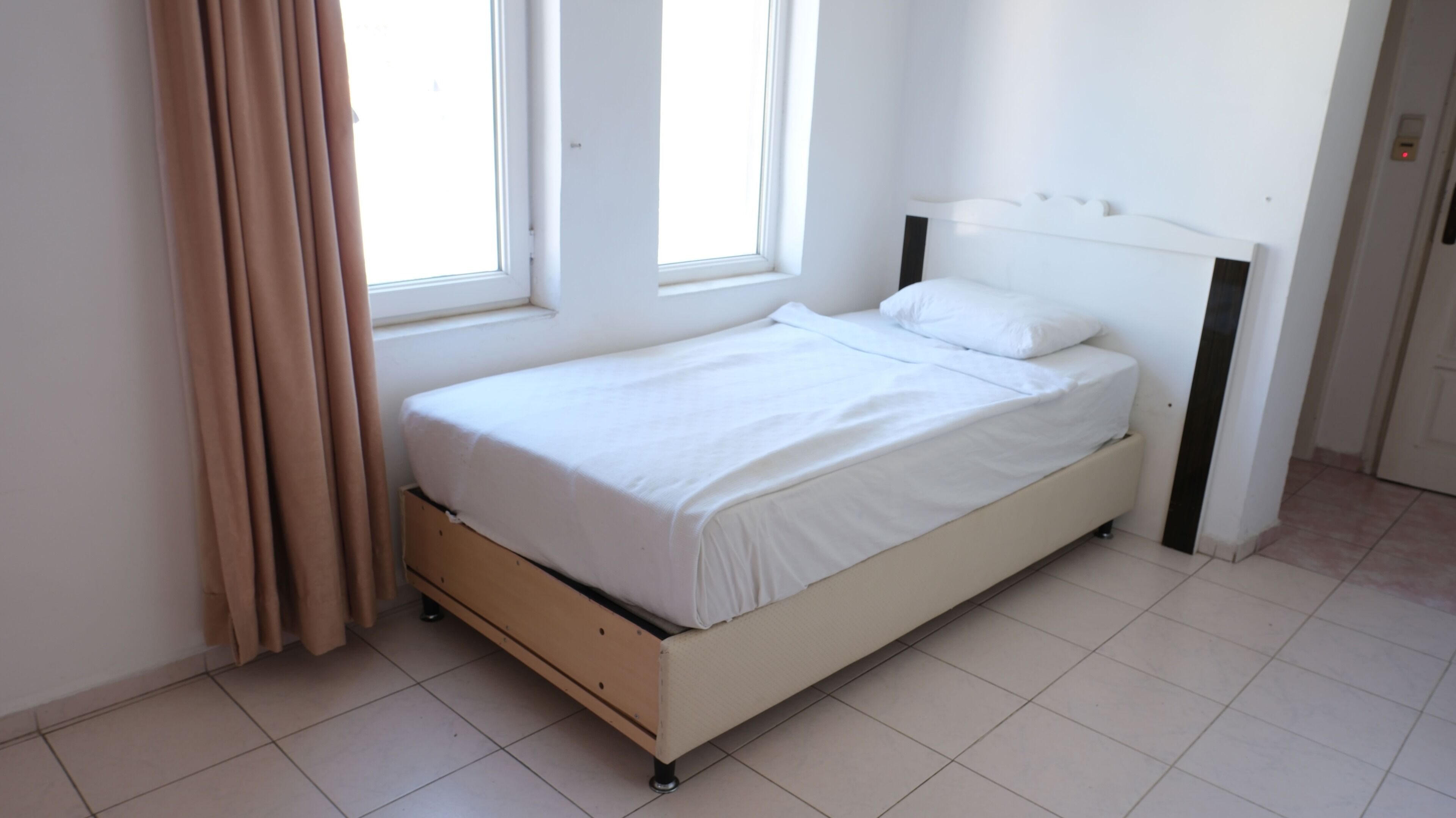 Una cama individual con colchón y cabecera blancos en una habitación con paredes blancas y suelo de baldosas.