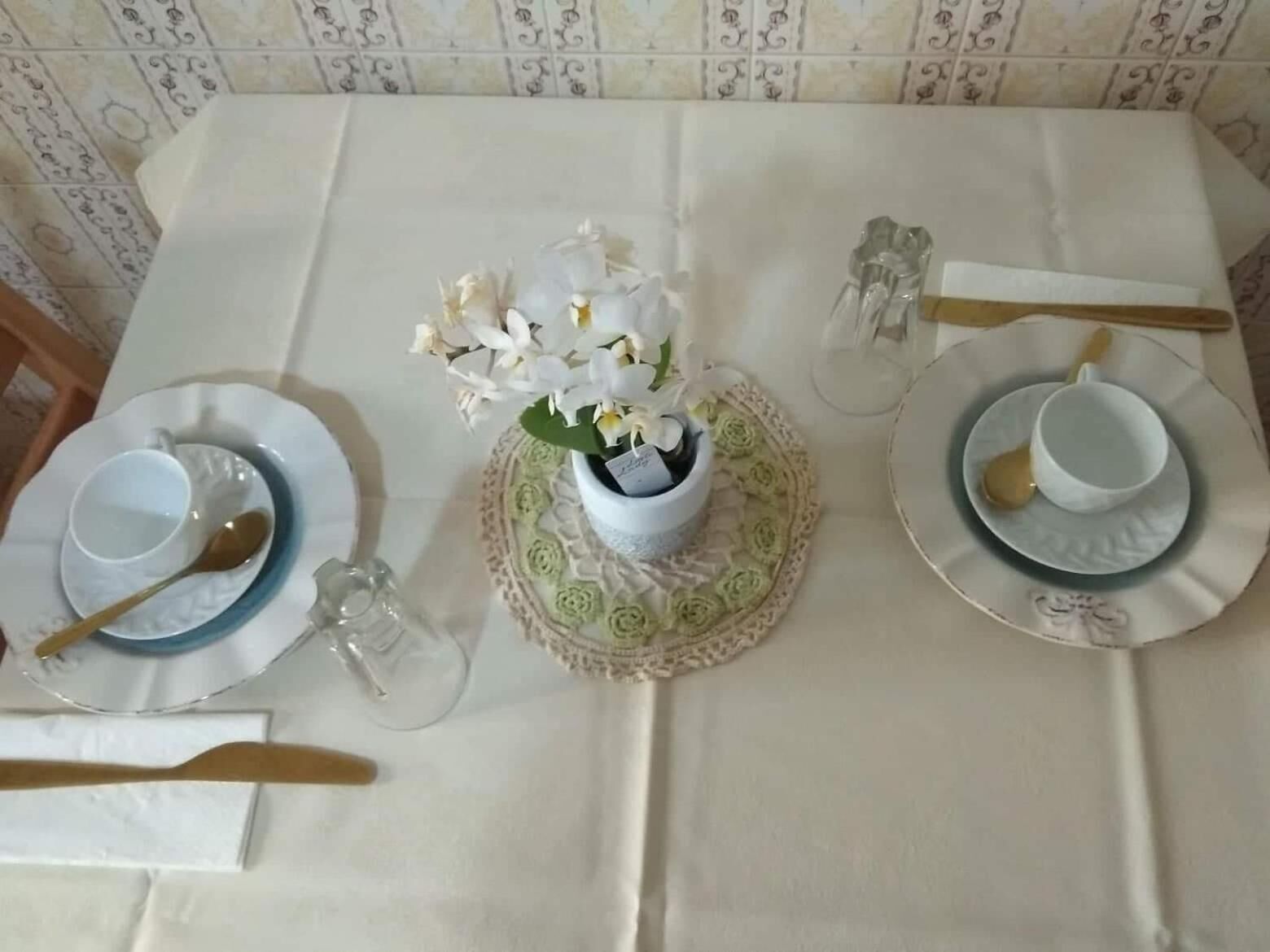 Una mesa de comedor impecable con mantel blanco, dos platos con cuencos, cucharas, una copa y un florero con flores blancas.