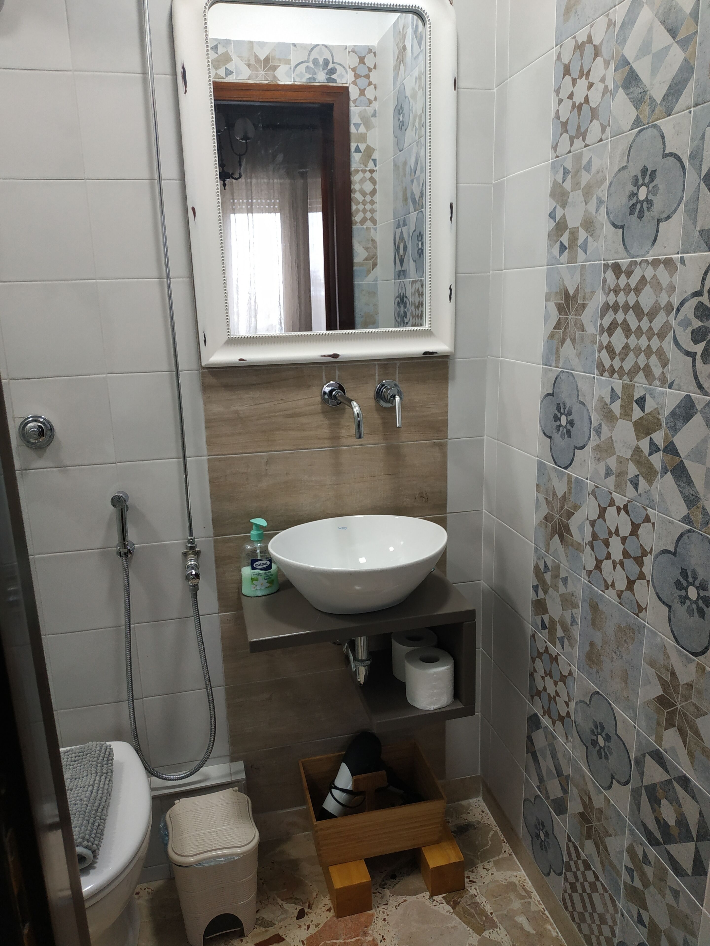 Ein Badezimmer mit einem weißen Waschbecken, einem Spiegel darüber, einer Dusche und einer Toilette. Auf dem Boden steht eine kleine Holzschachtel.