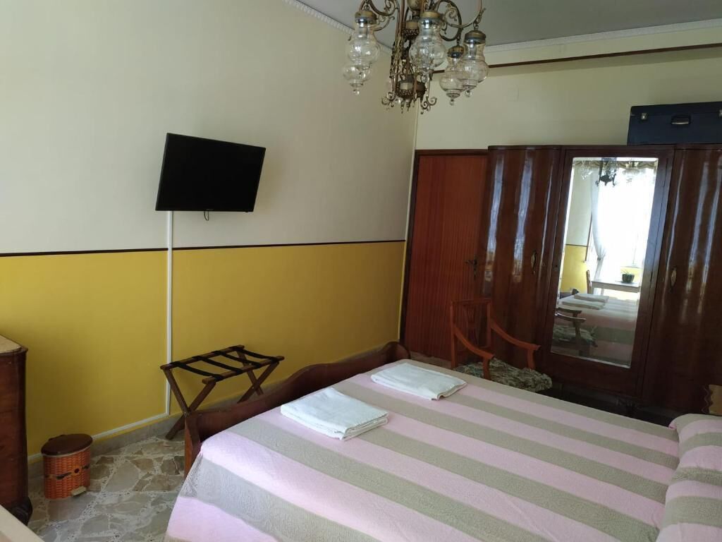 A hotel room with a bed, a television mounted on the wall, a wooden wardrobe, and a chandelier.