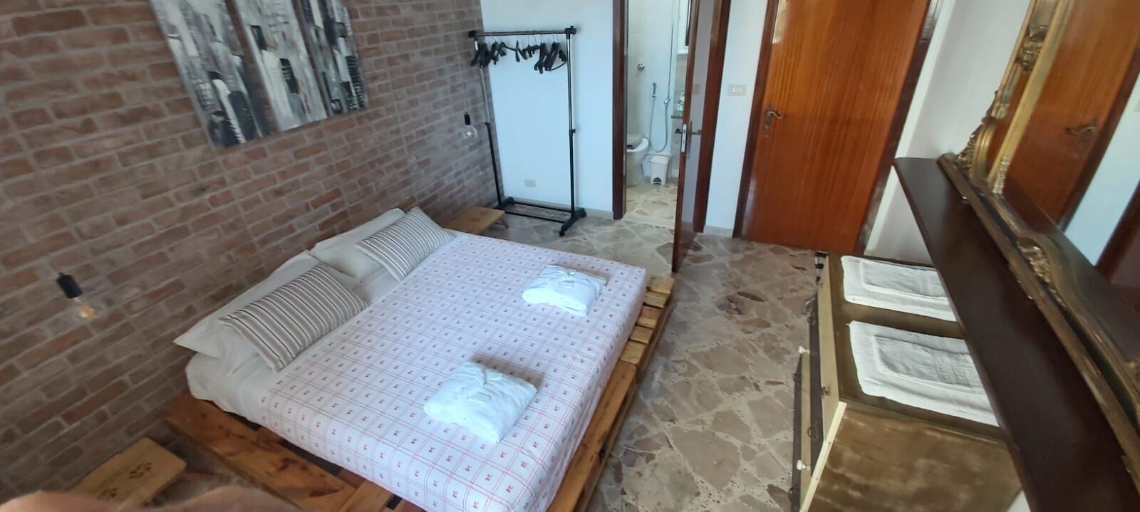 Une chambre avec un mur en briques, un lit recouvert d’une couvre-lit à motifs, une tête de lit en bois et une commode en bois.