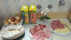 Una mesa de buffet con diversos alimentos, incluyendo bollos, zumos envasados, jamón en lonchas y patatas.