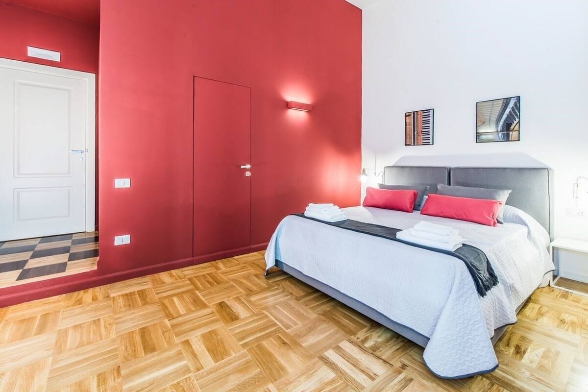 Un dormitorio con cama, pared roja, puerta blanca y suelo con patrón de espiga.