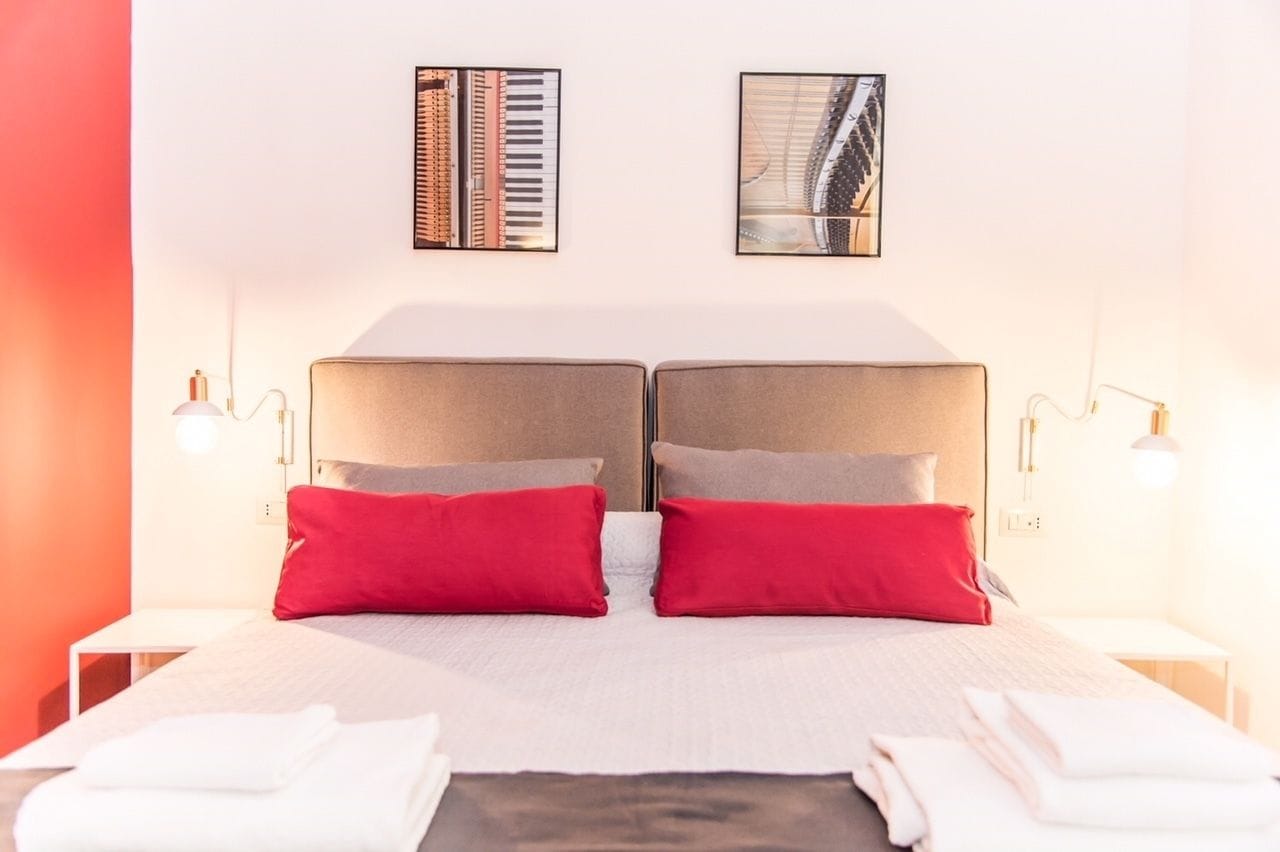 Cama doble con dos almohadas rojas, una colcha blanca y dos mesitas de noche con lámparas. Encima de la cama, hay dos cuadros enmarcados en la pared.