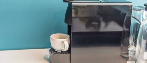 A black coffee machine with a white mug on its base, placed on a white surface against a teal wall.