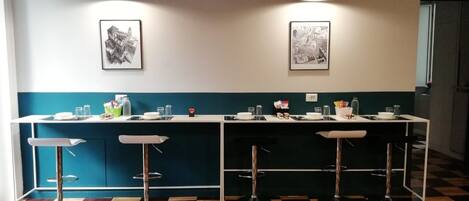 Una zona de bar con suelo a cuadros, taburetes, una barra con diversos objetos y dos luces fijadas a la pared.