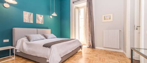 A bedroom with a bed, a nightstand, a window with curtains, a radiator, and a wall-mounted air conditioner.