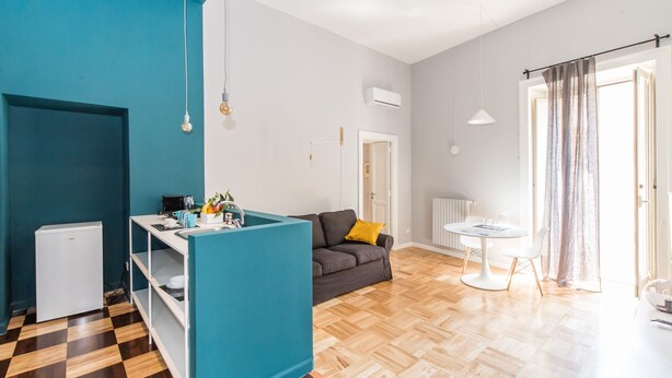 Een moderne woonkamer met een zwart-wit geruite vloer, een grijze bank, een blauwgroene scheidingswand, een kleine witte koelkast, een witte tafel met een stoel en een raam met gordijnen.