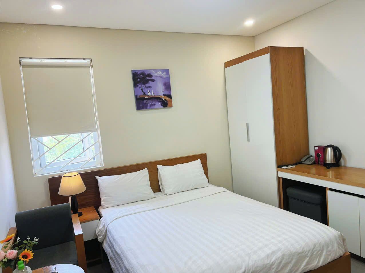 A hotel room with a bed, a desk, a chair, a lamp, a painting, and a wardrobe.