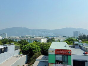 A building with a green roof, surrounded by trees and other buildings, with a distant view of mountains and industrial structures. - Anh Villa Hotel (Da Nang)