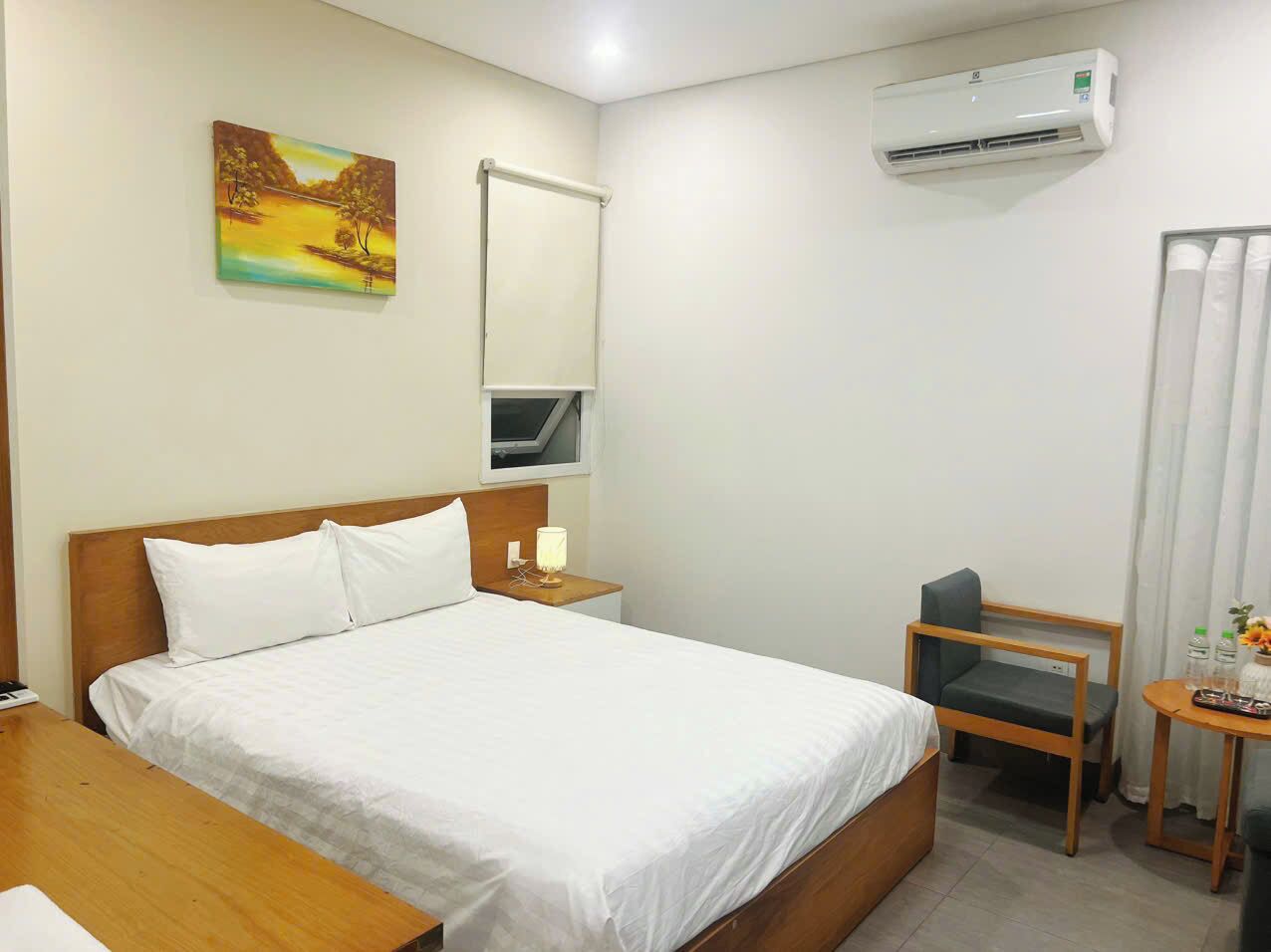 A hotel room with a bed, bedside table, chair, and an air conditioning unit.