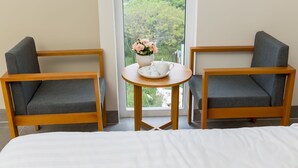A hotel room with two grey upholstered chairs, a small round wooden table with a white teapot and a bowl of pink flowers, and a large window offering a view of greenery. - Anh Villa Hotel (Da Nang)