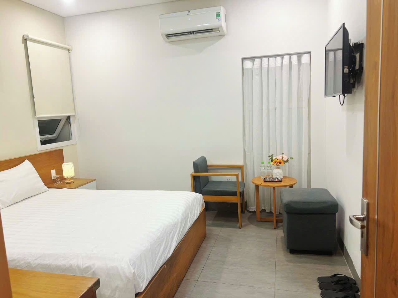 A hotel room with a bed, a desk, a chair, a small table with flowers, an air conditioning unit, and a television mounted on the wall.
