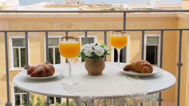 A balcony table with croissants and orange juice glasses, a vase with flowers, and a view of buildings in the background.