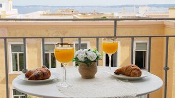 Een balkon tafel met croissants en glazen sinaasappelsap, een vaas met bloemen, en een uitzicht op gebouwen op de achtergrond.